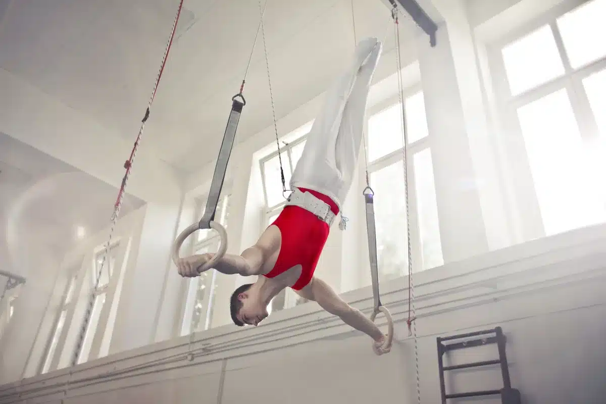 A young male gymnast in mid-routine on horizontal bar, muscles engaged, bar bending slightly, arena lights casting shadows across his concentrated face, hands gripping the apparatus with precision