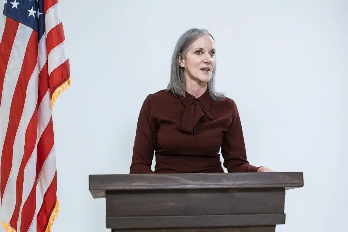 A professional woman in business attire standing at a podium with a South Carolina state flag in soft focus behind her, speaking to an unseen audience, determined expression, morning light through windows