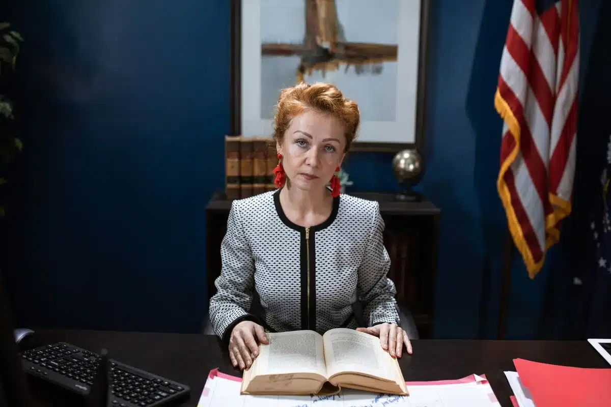 A woman in professional attire standing in front of an American flag, with formal Senate office lighting, her expression serious and focused.