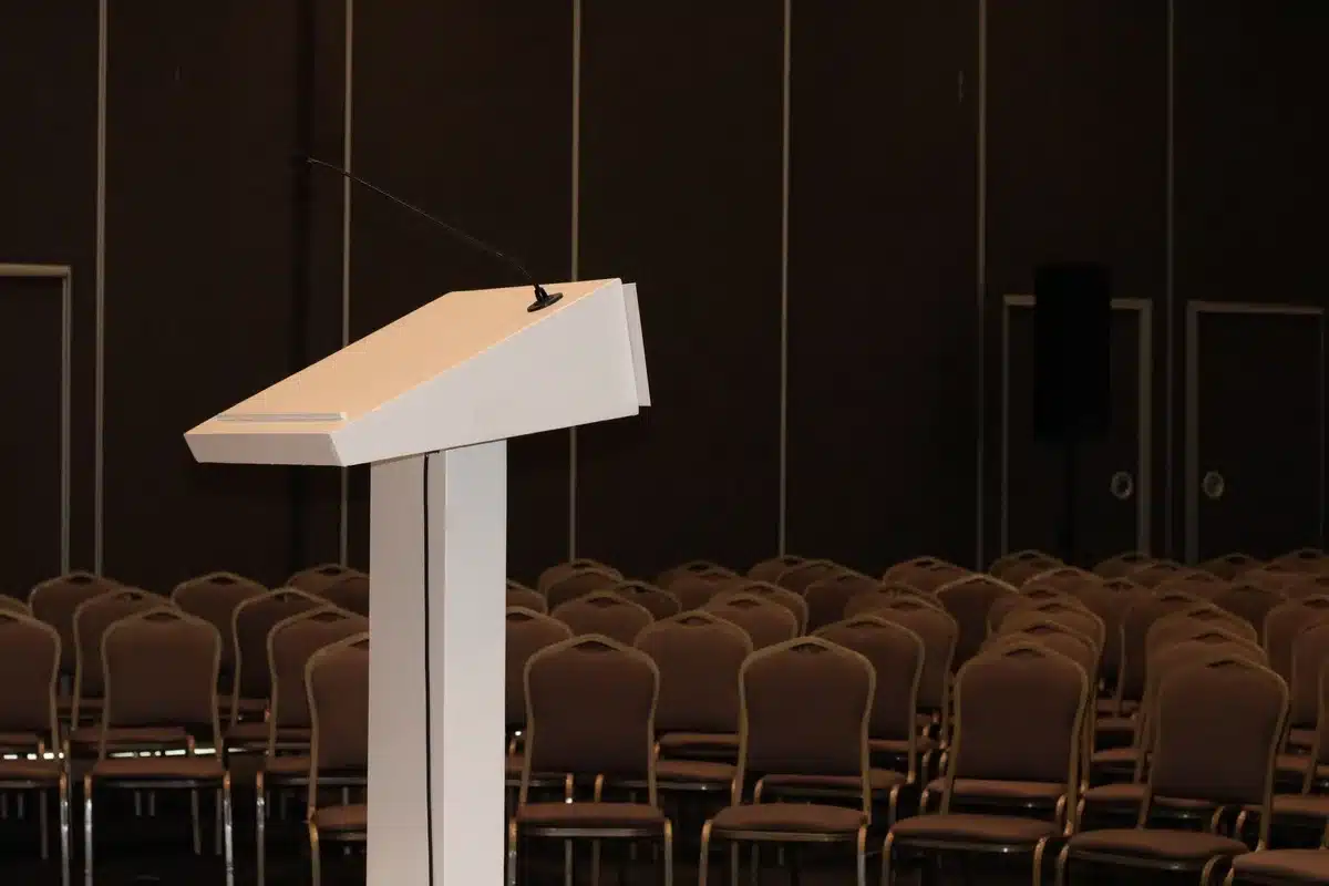 Podium and microphone in a dimly lit debate hall, empty chairs visible, cool blue and red lighting casting shadows across the stage, tension and political stakes evident