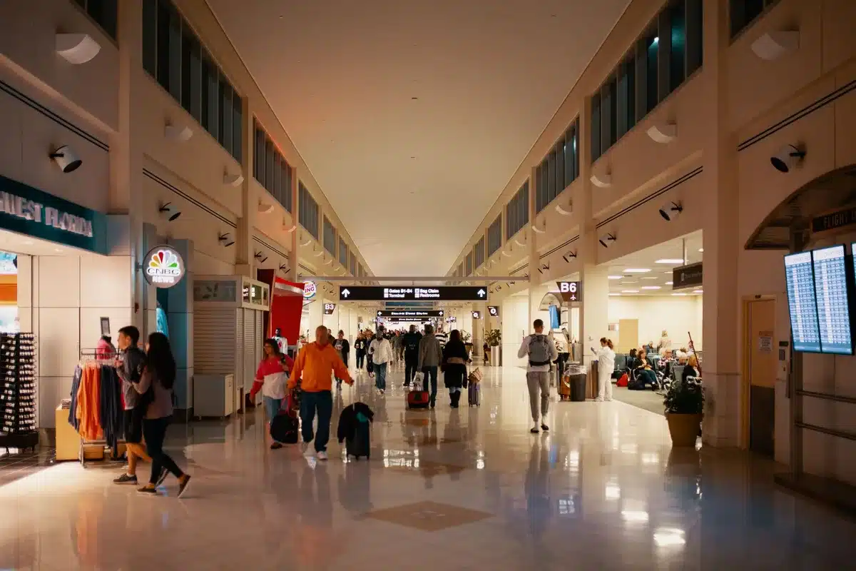 Passengers sitting in airport terminal with flight status boards showing cancellations and delays, some checking phones or looking at luggage.