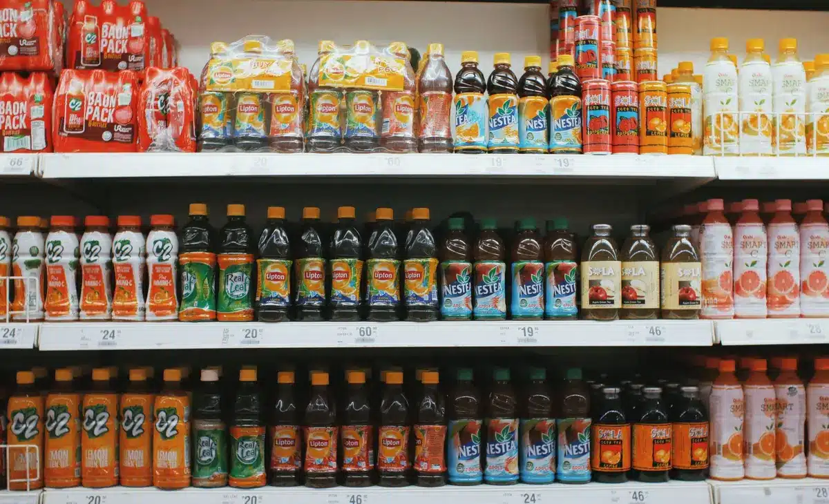 A grocery store aisle with price tags visible on shelves, shoppers selecting items, fluorescent lighting overhead.