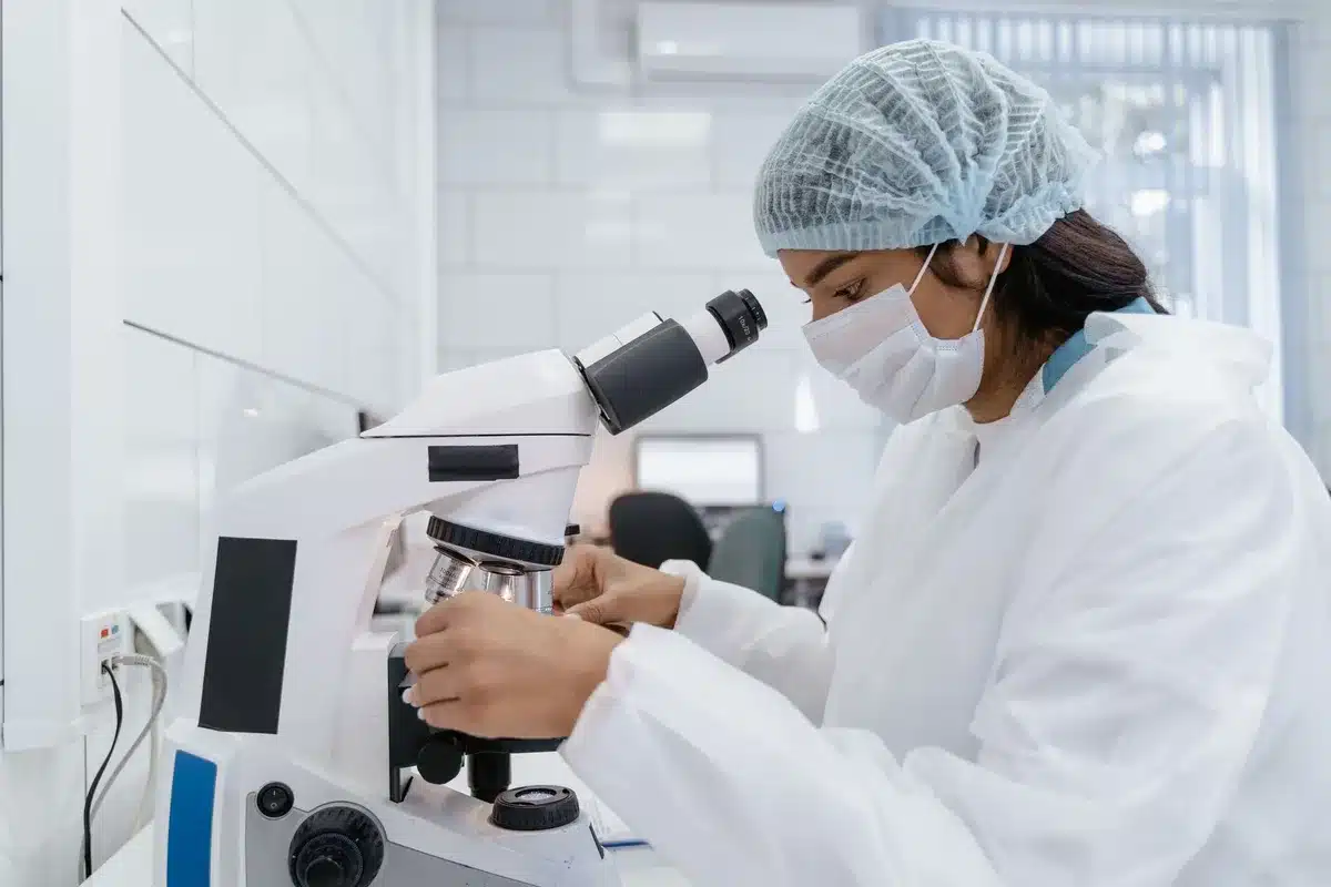 A researcher in a white lab coat examining a sample under a microscope with intense focus, hands positioned carefully on scientific instruments, soft laboratory lighting
