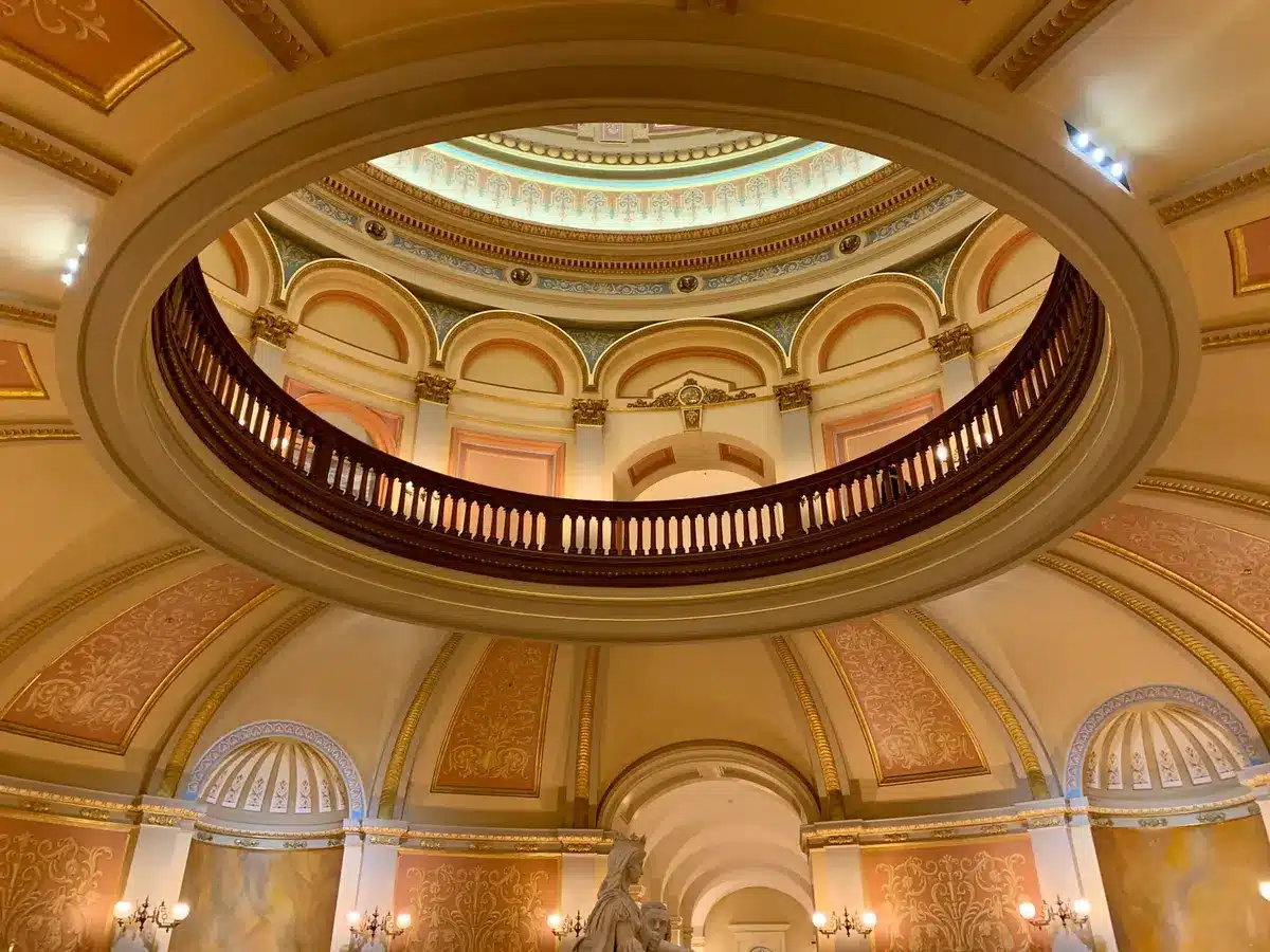 A Florida state capitol building exterior, columns and dome against a clear sky, empty plaza in foreground, afternoon light casting long shadows, symbolic of governmental authority and constitutional structure.