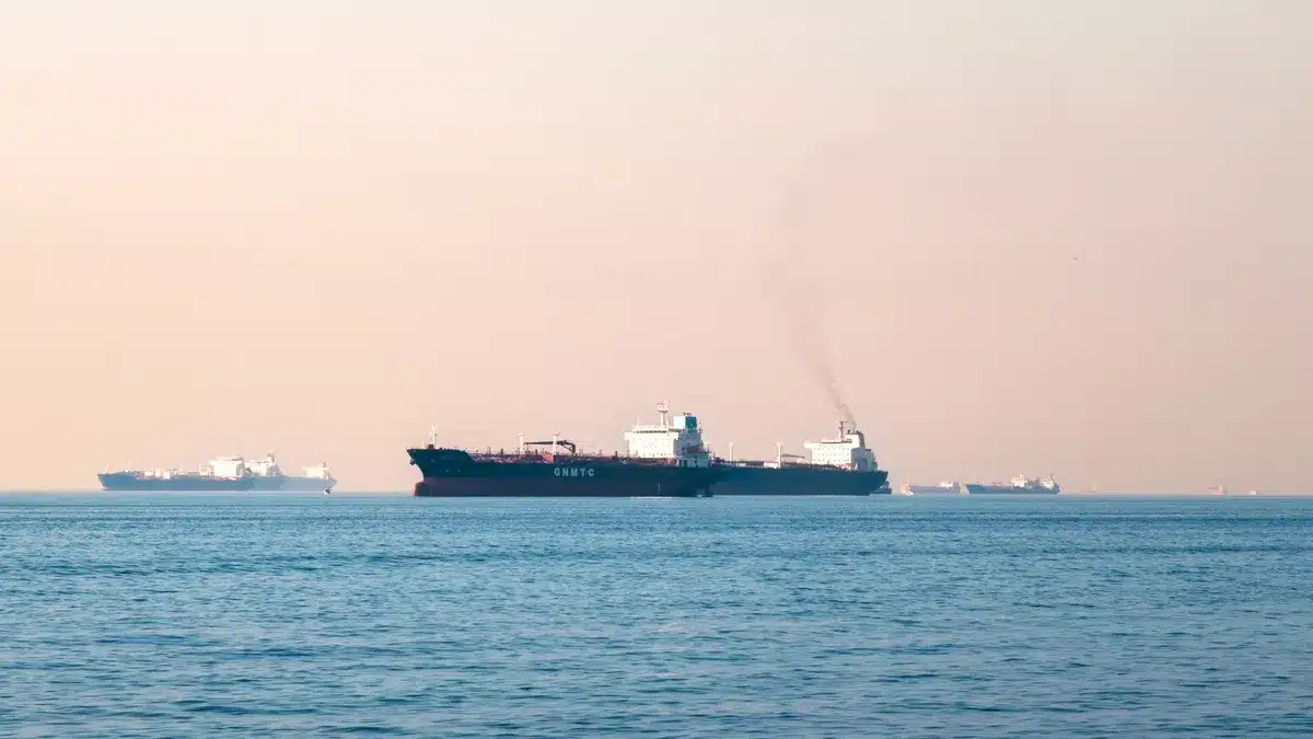 Tanker ship navigating narrow strait waters, rocky coastlines visible on both sides, hazy horizon, vessel silhouetted against pale sky