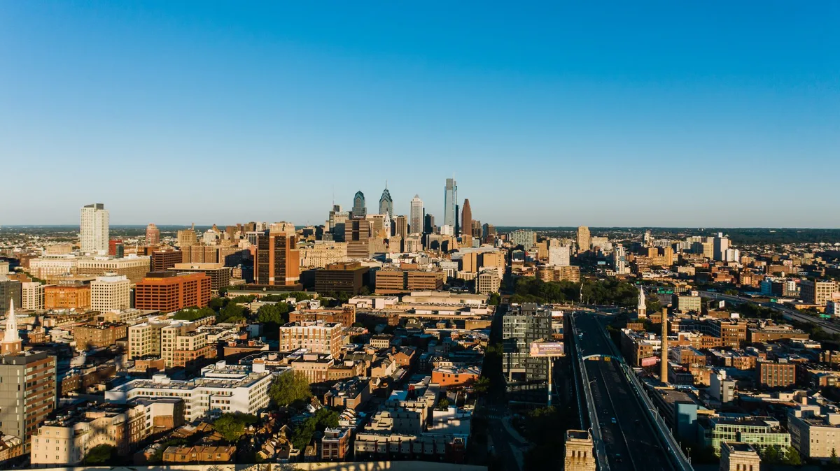 Three city skylines at sunset, each distinct&mdash;New England church steeples, Rocky Mountain peaks, historic Philadelphia architecture&mdash;representing the competing finalist cities for a major national gathering.