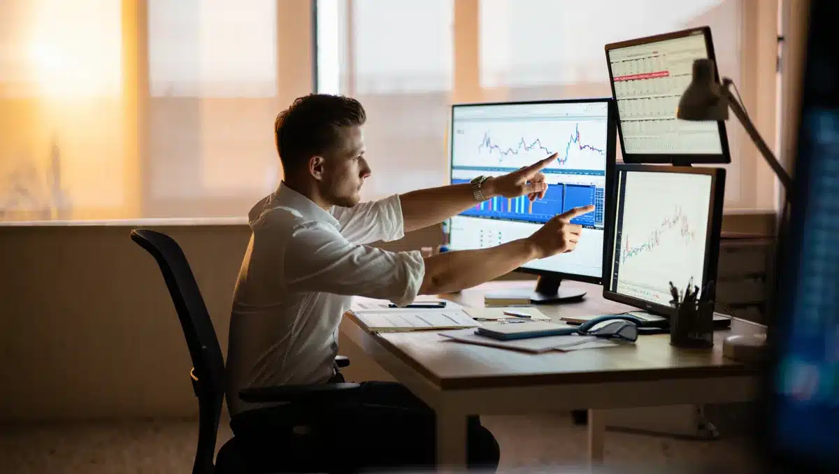 A financial analyst reviewing charts at a desk with multiple screens showing market data, hands pointing to rising trend lines, early morning light through office windows, papers with quarterly earnings reports visible — insight.