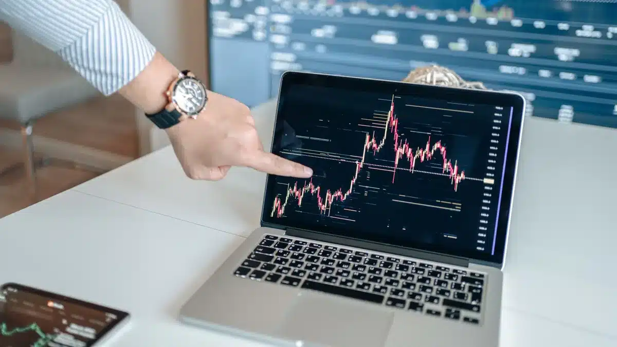 A trader's hand reaching toward a downward-pointing arrow on a glass surface, fingers mid-gesture above a glowing market dashboard, silhouette against blue and red trading screens, tension visible in body language.