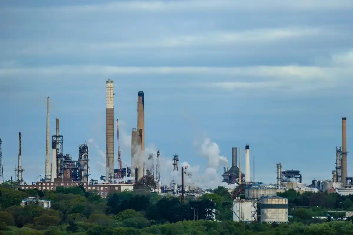 Oil refinery at night with illuminated storage tanks and pipeline infrastructure, flames visible from flare stacks against dark sky, industrial complexity and energy production scale