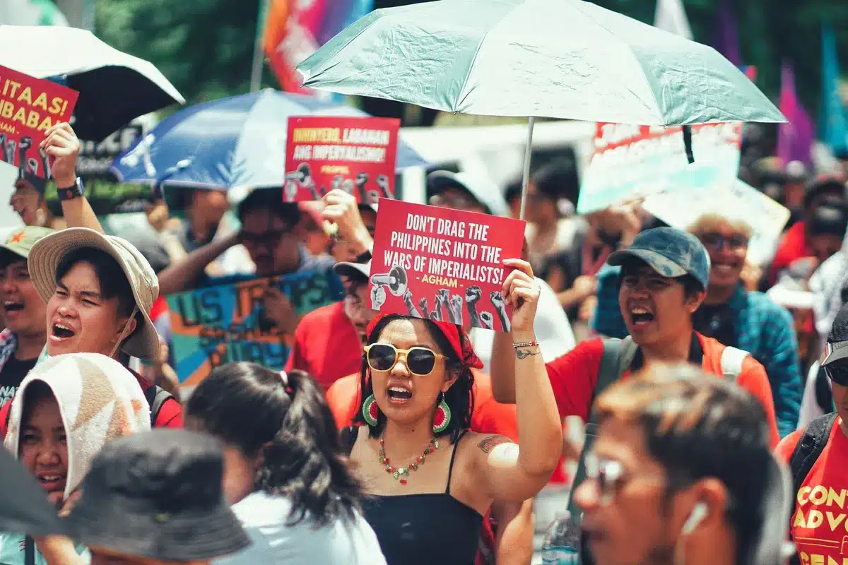 A crowded political rally with diverse supporters holding campaign signs and banners, captured from above showing the scale of grassroots mobilization, faces obscured in motion, energy and momentum visible