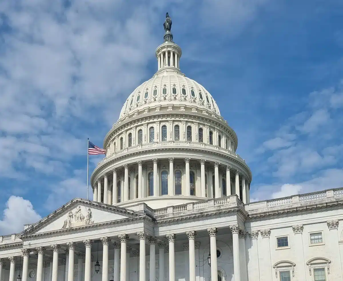 A modern congressional chamber with empty marble columns and ornate railings, symbolic of institutional power and democratic processes. Sunlight streams through tall windows, casting long shadows across polished floors, evoking both grandeur and the weight of structural change.