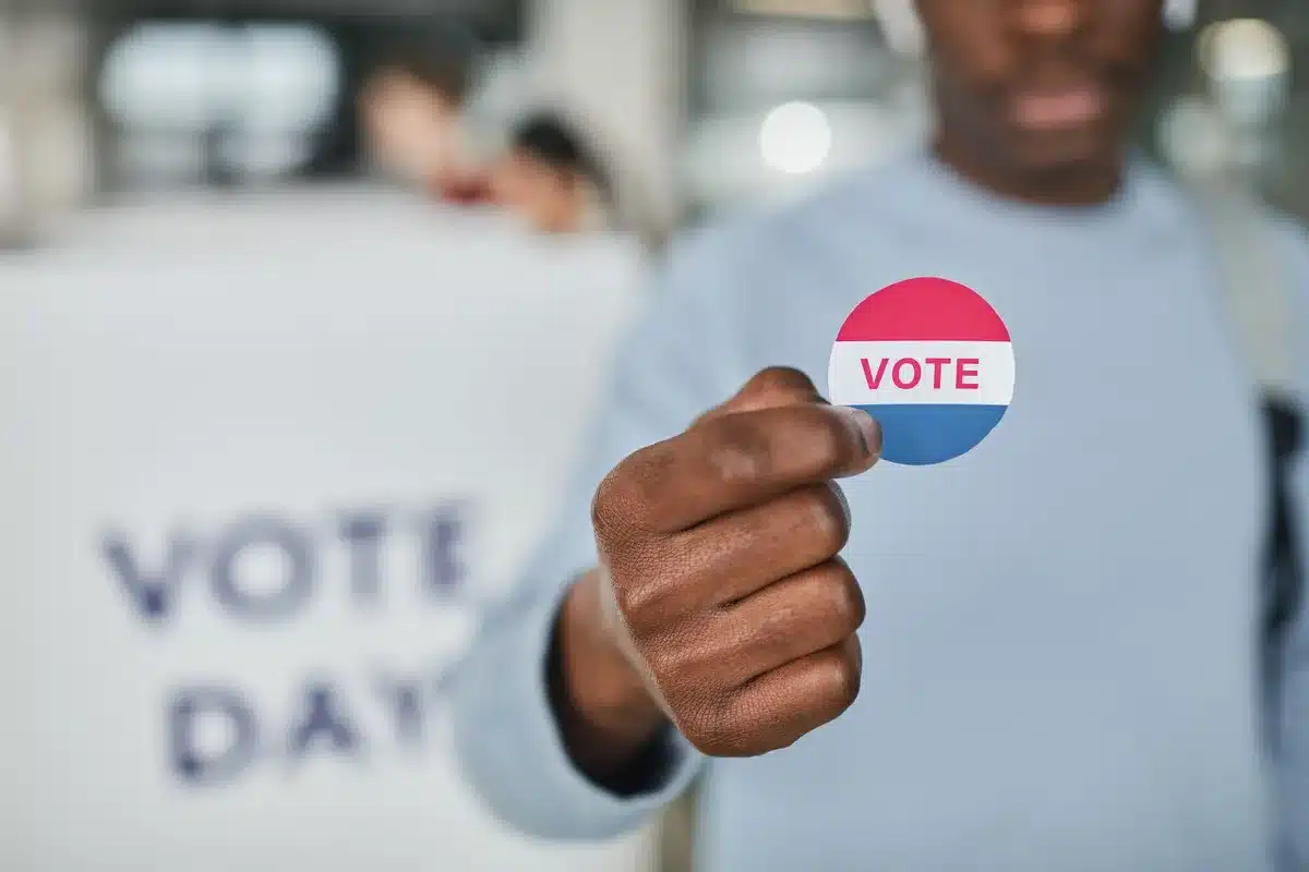 A diverse voting place with citizens at ballot stations, poll workers assisting voters, American flag visible in the background, civic participation and democracy in action.