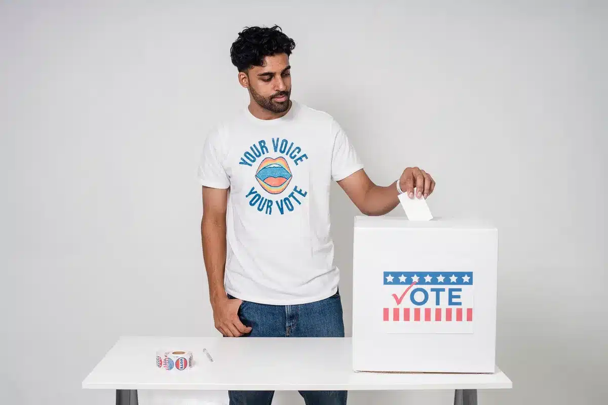 An empty voting booth with voting machines visible, a single ballot or pen on the table, fluorescent lighting overhead, a symbol of electoral choice and democratic process.