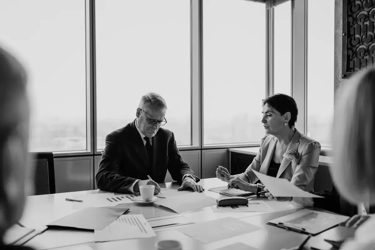 A modern office boardroom with large windows overlooking a city skyline, a wooden conference table with chairs, and contract documents spread across the surface, illuminated by soft afternoon light, suggesting corporate negotiation and legal commitment