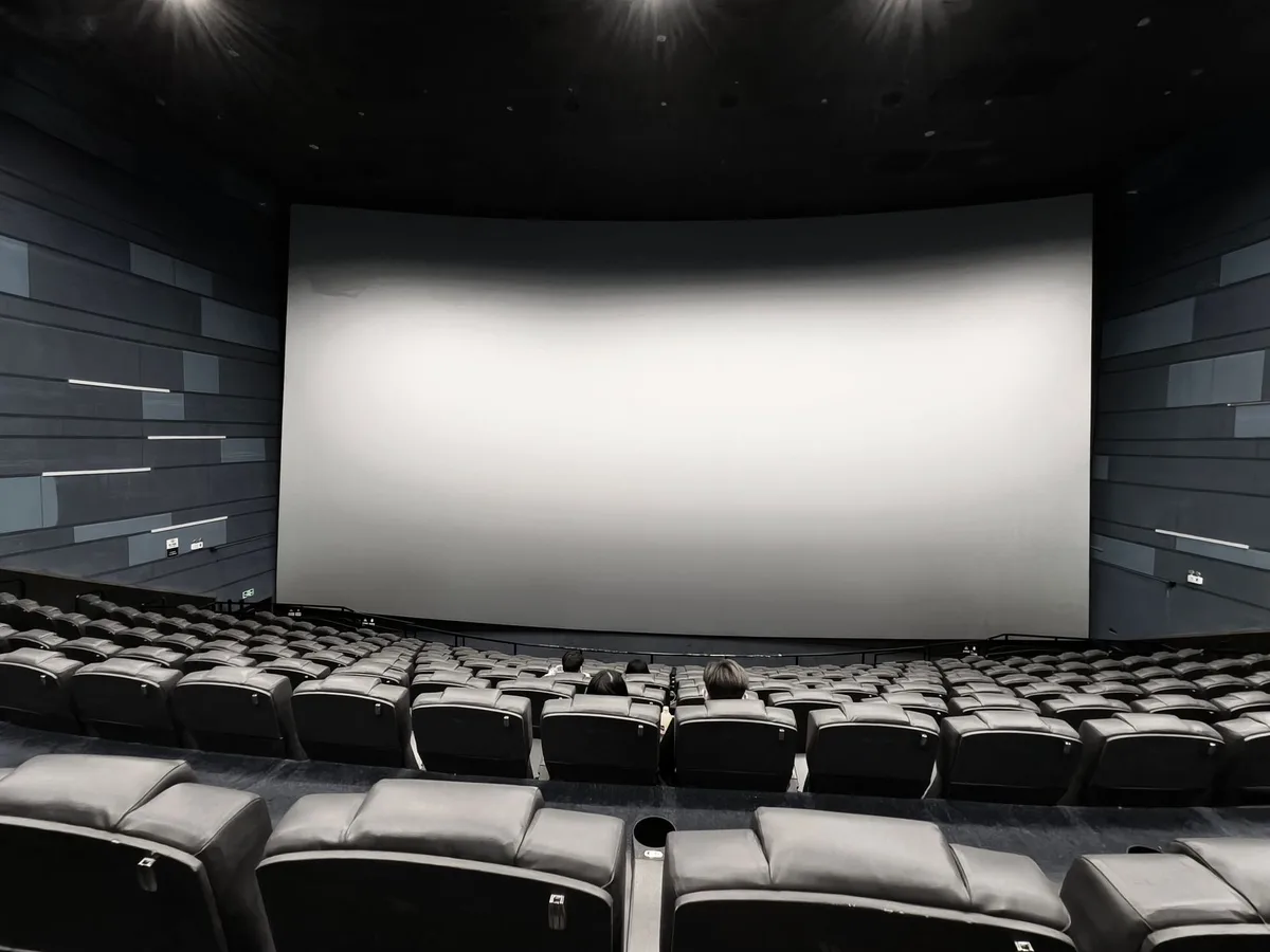 A movie theater interior at dusk with rows of empty seats facing a dark screen, subtle red and blue lighting from exit signs, and a single spotlight illuminating the projection booth, evoking anticipation and the theatrical experience at stake