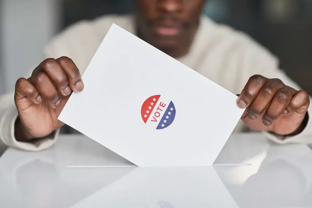 A voting booth setup in a Minnesota gymnasium or community center, with an American flag in soft focus behind it, voters entering to cast ballots