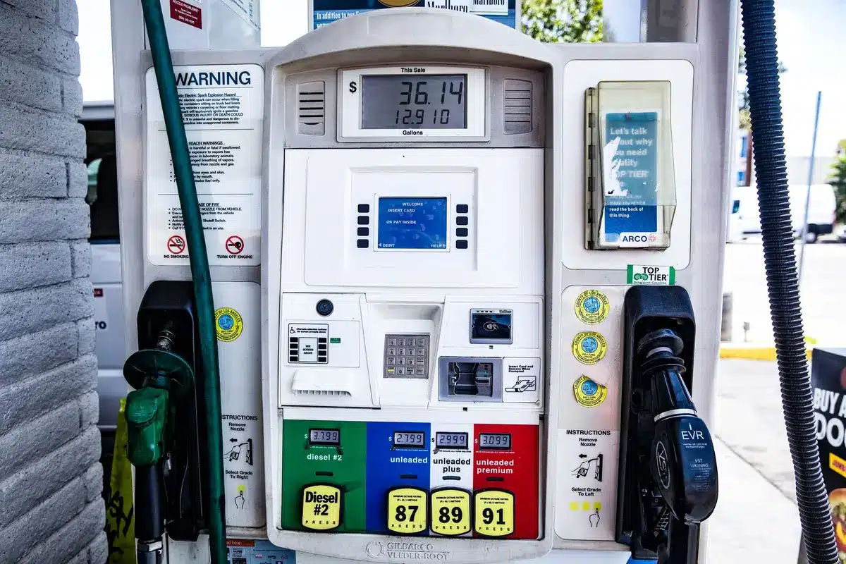 Gas station pump display showing high prices, numbers glowing in red against dark background, a single customer's hand reaching toward the pump handle in shadow