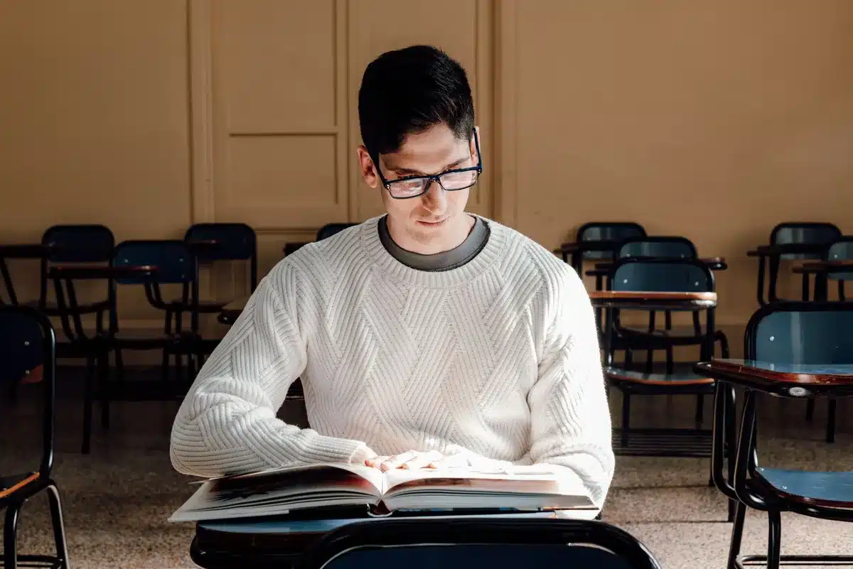 Graduate student studying at a desk with textbooks and laptop, late afternoon light streaming through a window, contemplative expression