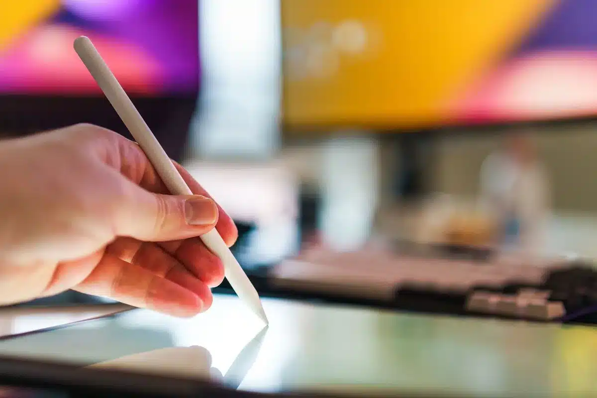 Close-up of a designer's hands on a keyboard and trackpad, illuminated by a glowing monitor displaying design tools and interface elements, evoking focused creative work amid uncertainty