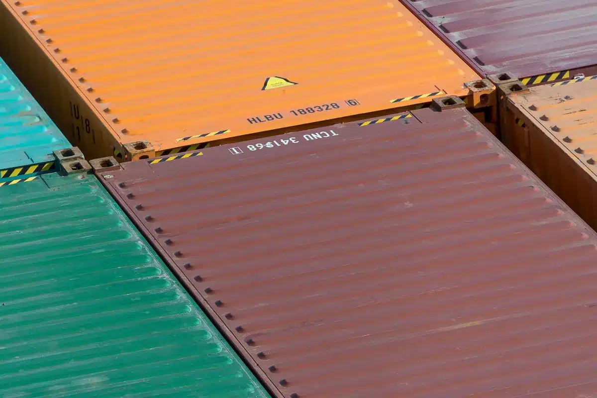 Empty shipping containers stacked at a busy port terminal, cranes and cargo ships visible in the background, sunlit industrial waterfront landscape conveying trade disruption.