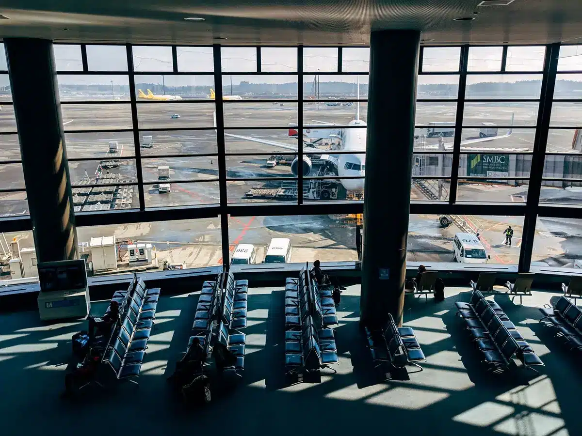 A crowded airport terminal with passengers waiting at departure gates, some standing near flight information boards displaying cancellations and delays, overhead lighting casting harsh shadows, the scene conveying frustration and disruption