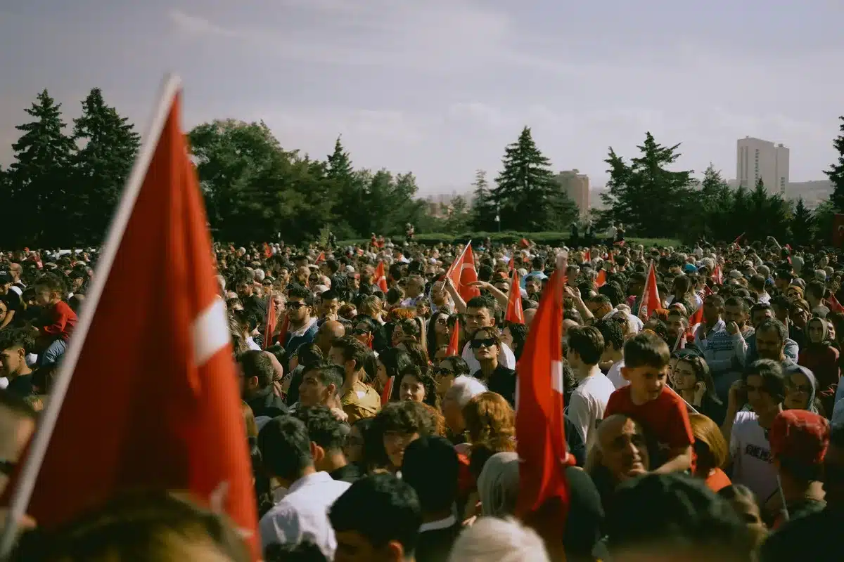 A gymnasium filled with young supporters, many standing with raised fists and cheering, stage lights illuminating the crowd, a podium with campaign signage visible, energy and collective enthusiasm filling the space