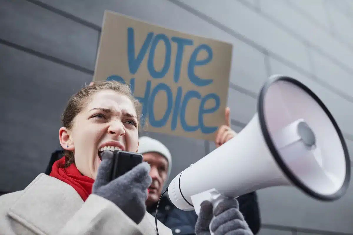 Young first-time voters at a rally entrance, holding campaign literature and merchandise, faces animated with hope and engagement, sunlight streaming through gymnasium doors, a sense of civic participation and youthful energy