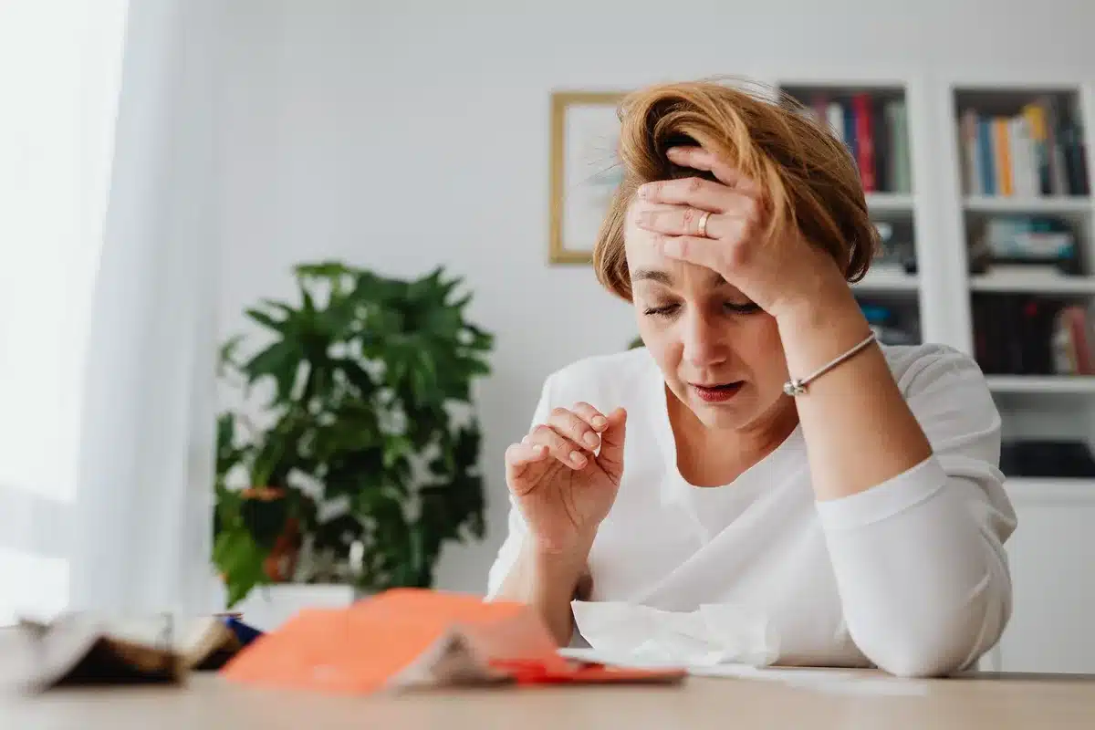 Middle-aged person sitting at a desk, head in hands, papers and bills scattered, morning light through window, household budget visible