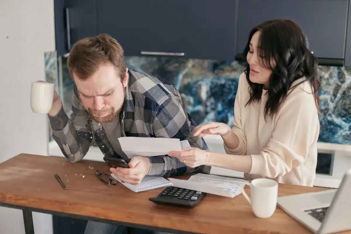 A middle-aged person sitting at a desk surrounded by bills and financial documents, head in hands, looking worried. Warm indoor lighting casts shadows across stacks of papers and a calculator.