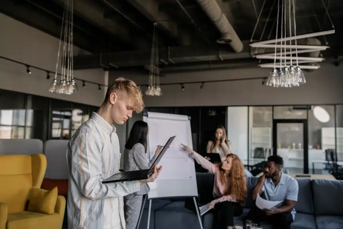 Young professionals in an open office workspace, seated at desks with laptops, some in conversation, natural daylight streaming through large windows, focused and collaborative atmosphere
