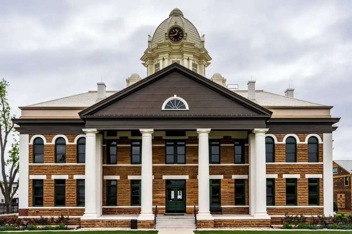 A federal courthouse entrance with columns and flags, morning light casting long shadows across stone steps, emphasizing institutional solemnity and legal weight