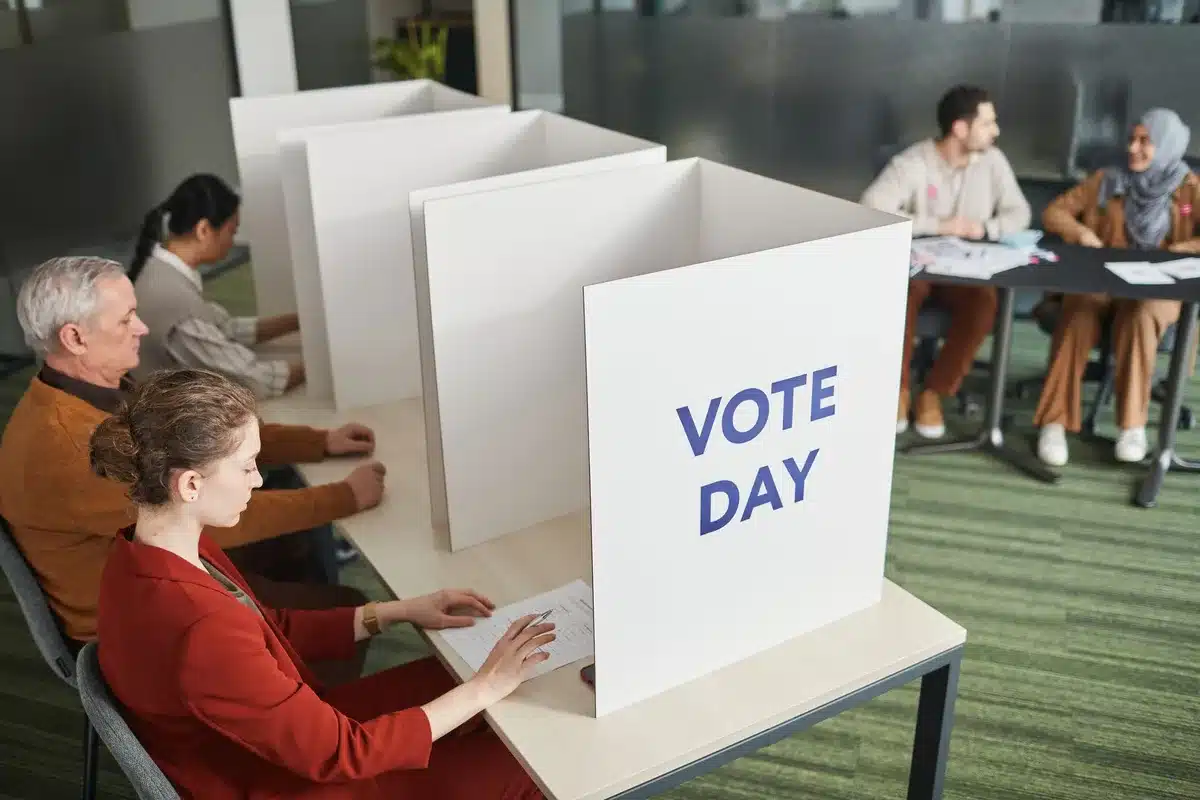 A diverse crowd of anonymous voters casting ballots at polling stations, some studying sample ballots, others feeding ballots into machines, fluorescent light casting shadows on voting booths and election signage in the background, anticipation and civic engagement visible