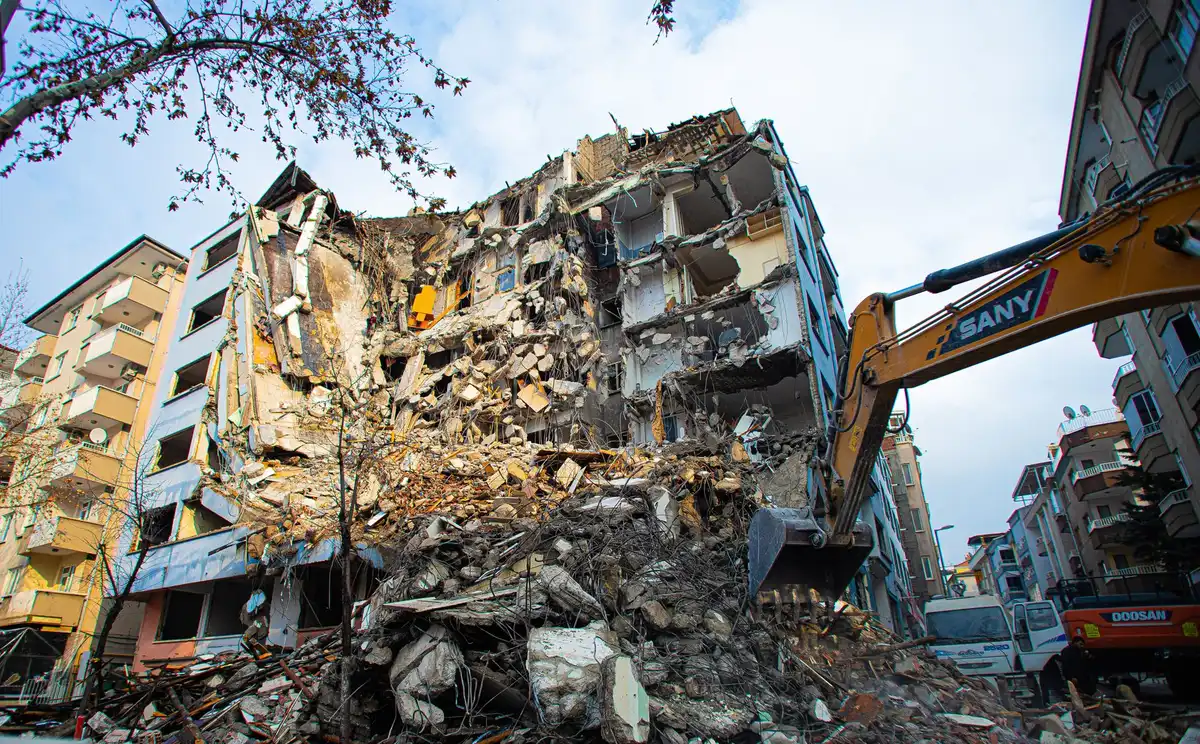 A collapsed building facade amid urban debris, rescue workers in hard hats moving through rubble, emergency vehicles and yellow caution tape visible in the background, dawn light casting long shadows across the destruction scene