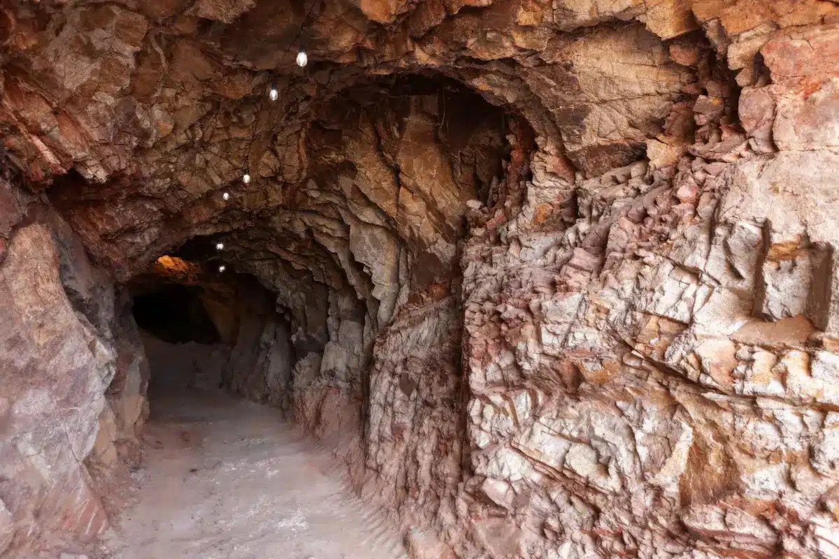 An underground mine tunnel with exposed ore veins glinting under work lights, deep within the earth where precious metals lie dormant, the scale of geological time and resource wealth visible in layered rock.