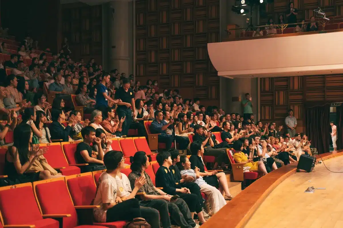 A packed historic opera house with an audience watching two candidates standing at debate podiums, one woman and one man, under bright stage lights. Audience members visible in the foreground show mixed reactions.
