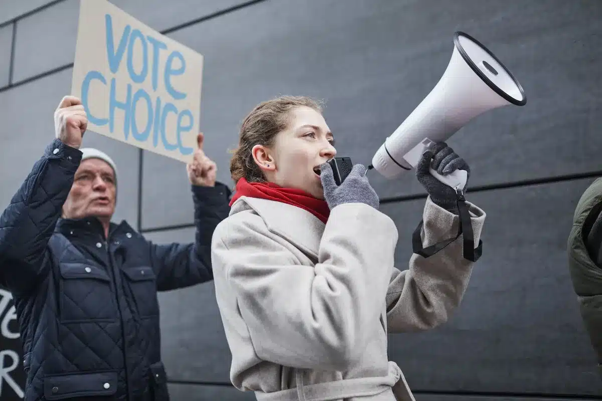 An appointed interim senator at a campaign podium in a South Carolina venue, addressing supporters with American flags visible in the background, determination visible in the moment