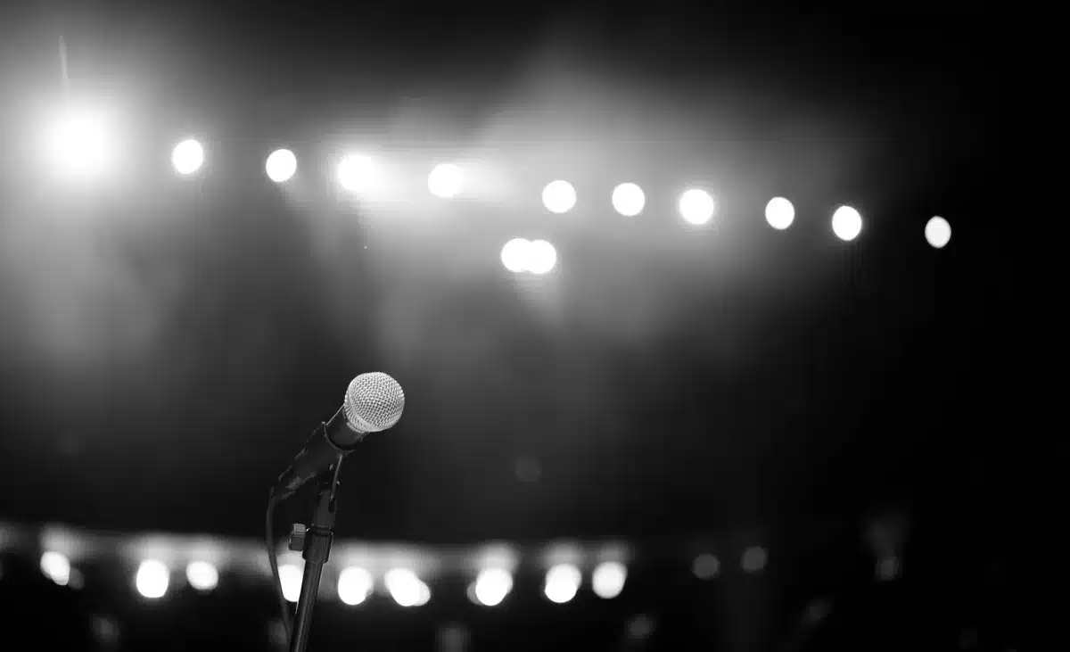 A spotlight shining on an empty stage with a single microphone stand, dramatic shadows across wooden floorboards, dust particles visible in the beam, a coiled cable trailing into darkness