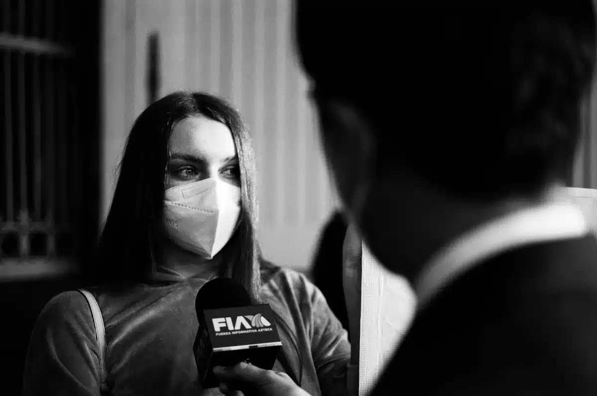 A female journalist's silhouette facing away from a microphone in a dim press room, hands raised in question, tension visible in posture.