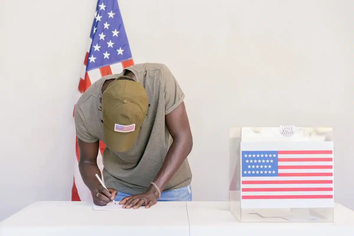 Democratic primary voting booth with diverse hands reaching toward ballots, fluorescent polling place lighting, faces blurred or turned away