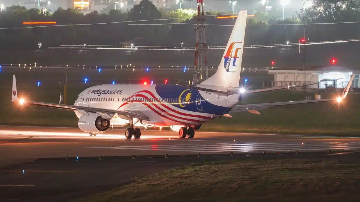 An empty nighttime runway at an international airport with runway lights illuminated, taxiway markings visible, and a control tower silhouette in the distance.