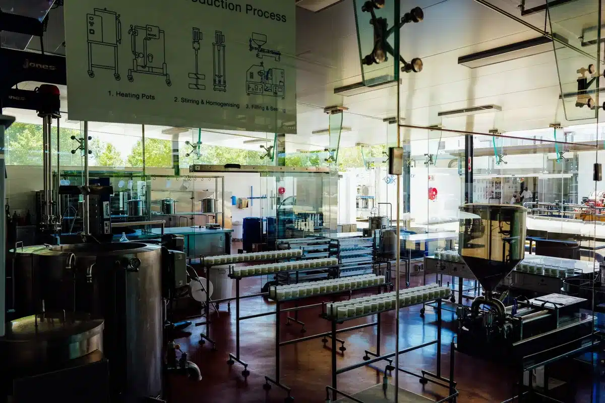 A wide view of a manufacturing plant floor with military equipment in assembly stages, workers in safety gear moving through the space, natural light streaming through industrial windows