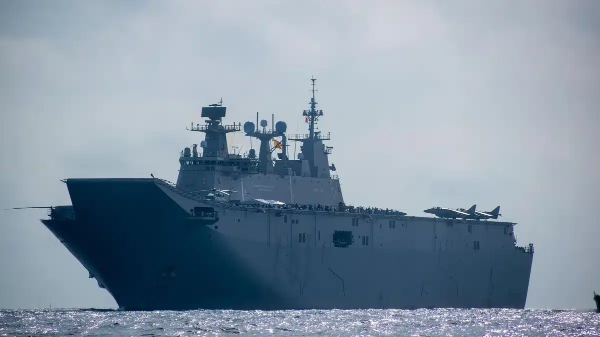 A gray aircraft carrier cutting through blue ocean waters, sailors visible on the flight deck under bright sunlight, the ship's superstructure rising against a clear horizon, isolation and vastness conveyed.