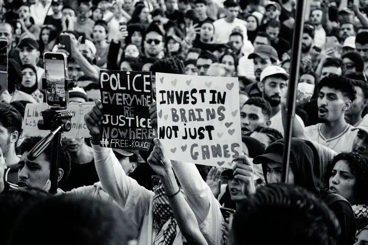 Campaign rally or town hall with mixed-age crowd, some holding signs related to economic reform or healthcare, energy and engagement visible