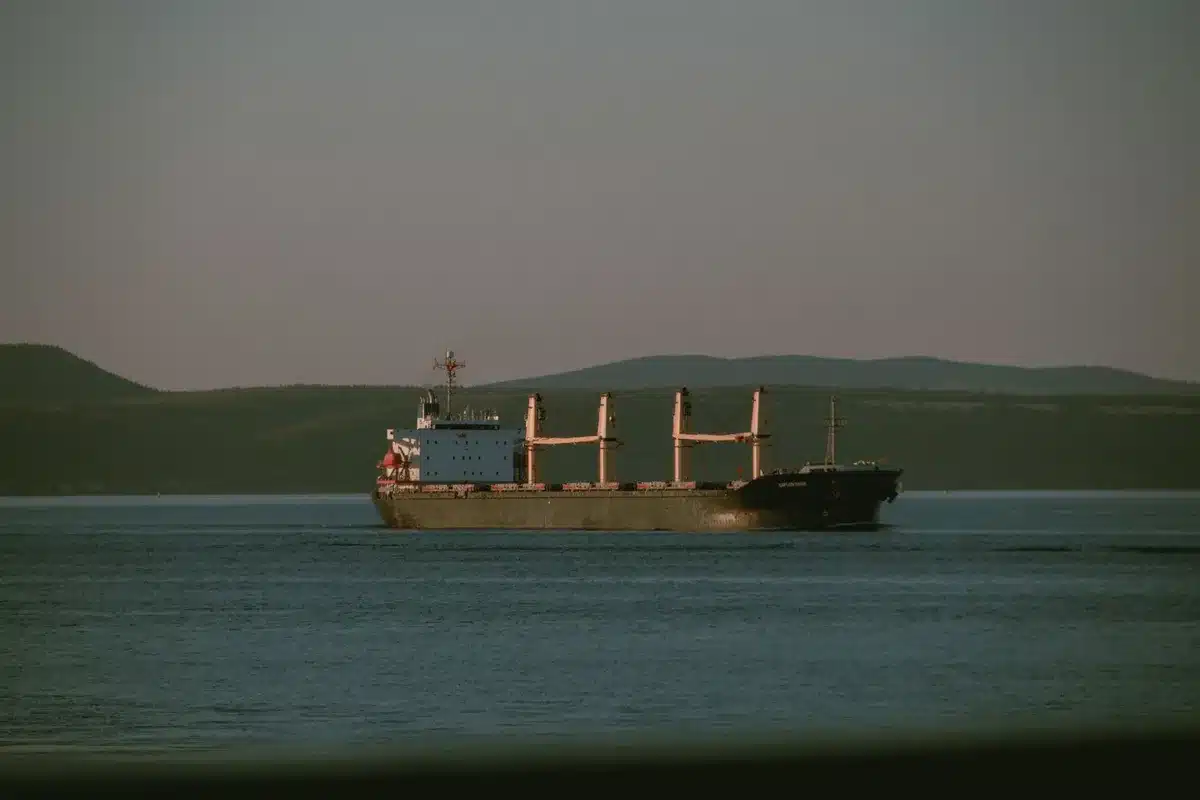 An empty shipping lane with a single cargo vessel silhouetted against calm waters, surveillance radar equipment visible on a distant platform, morning light breaking through haze, the quiet tension of a contested waterway