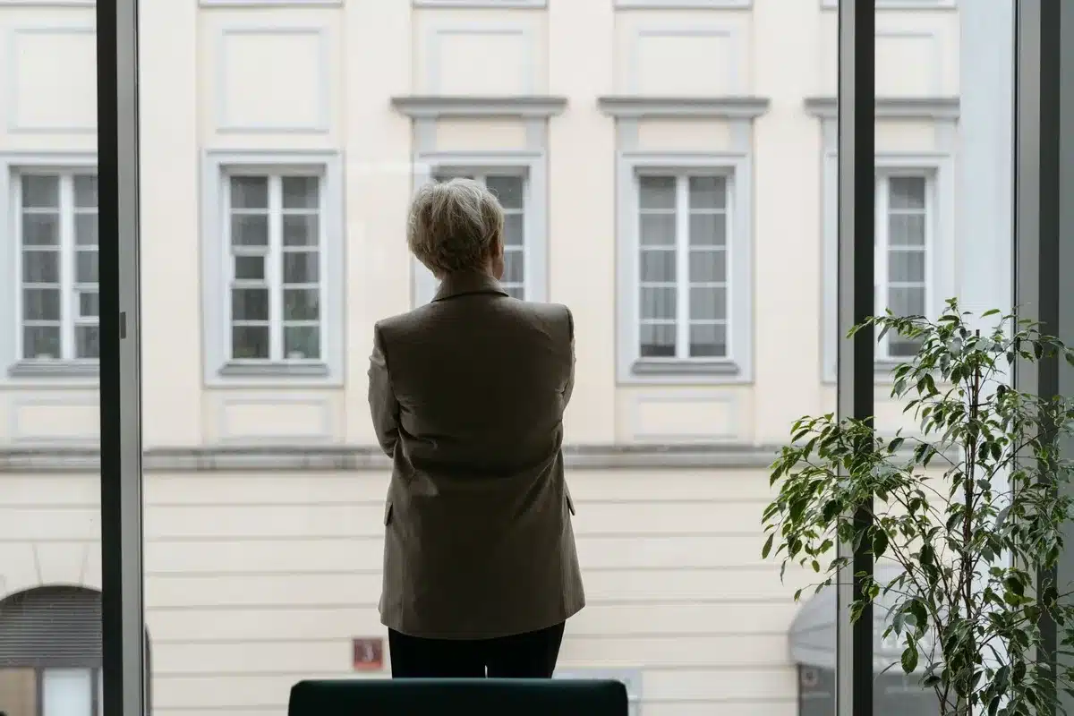 Anonymous man in contemplative pose, hands clasped, facing away from camera toward a window with diffused light, muted tones, introspective mood