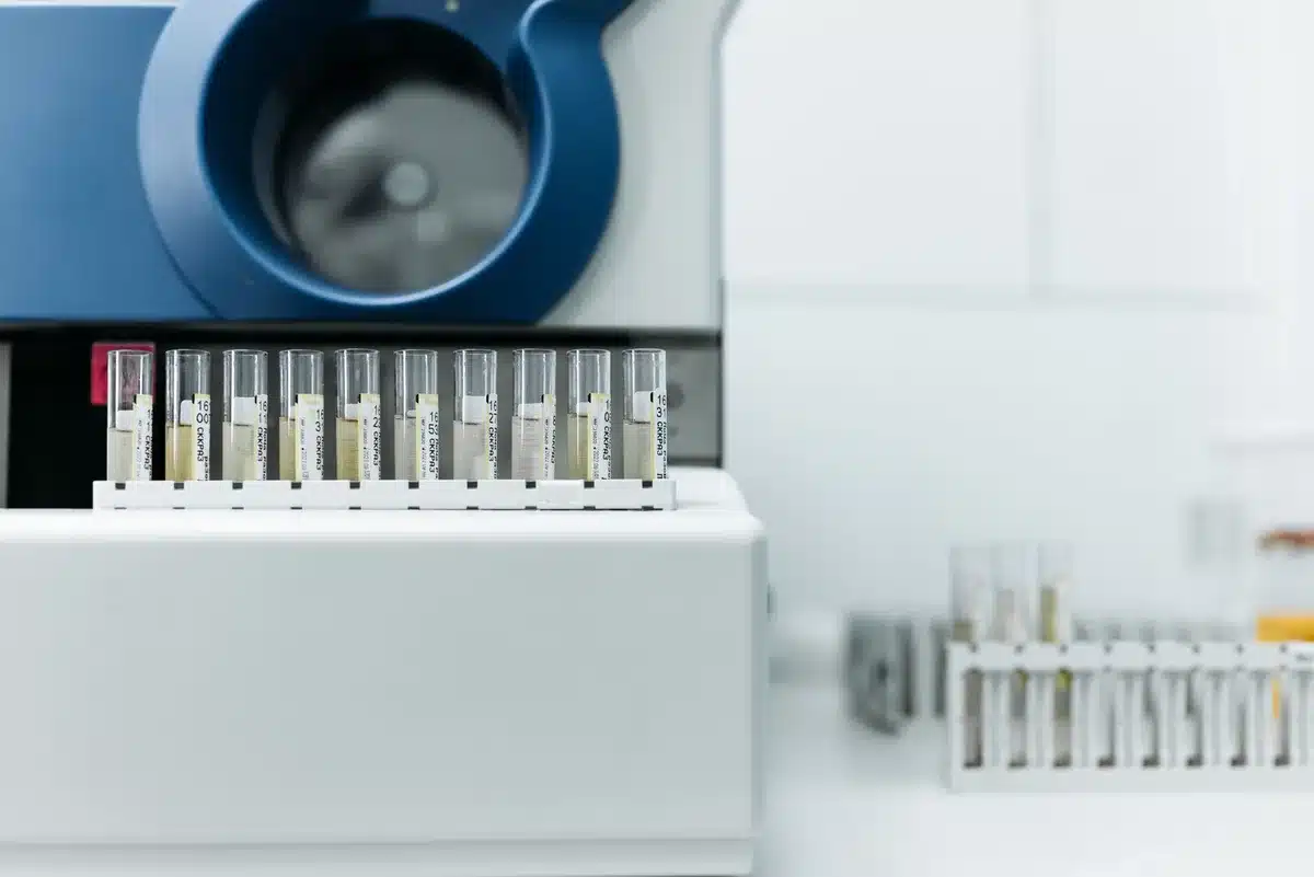 A federal laboratory workstation with DNA sample vials and testing equipment arranged on a sterile counter, harsh white lighting illuminating test tubes and biometric data sheets, a computer screen displaying genetic profile information in the background.