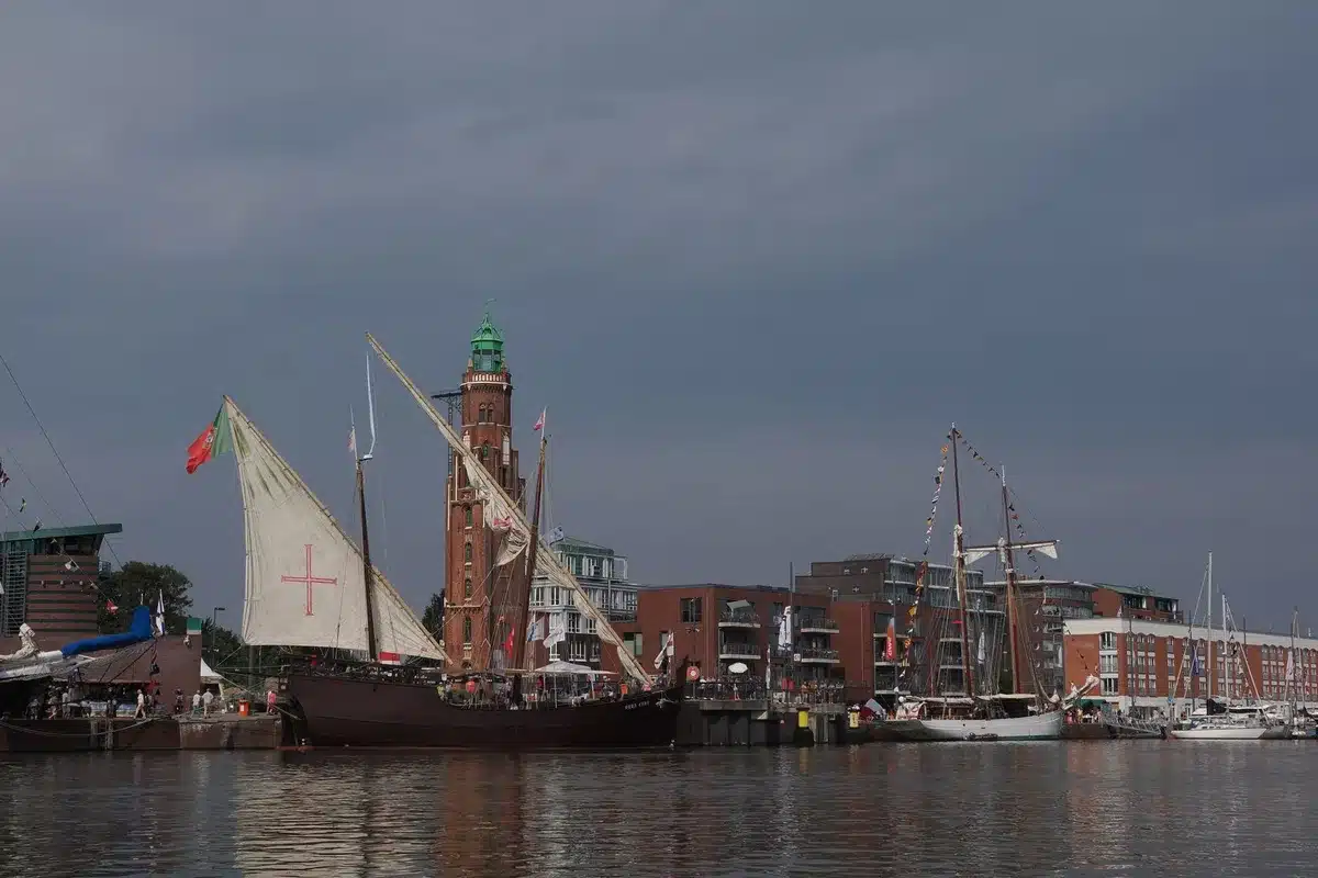 Majestic tall ships with billowing sails moving through a harbor under blue skies, passing an iconic statue in the background, international flags flying from masts, crowds lining waterfront