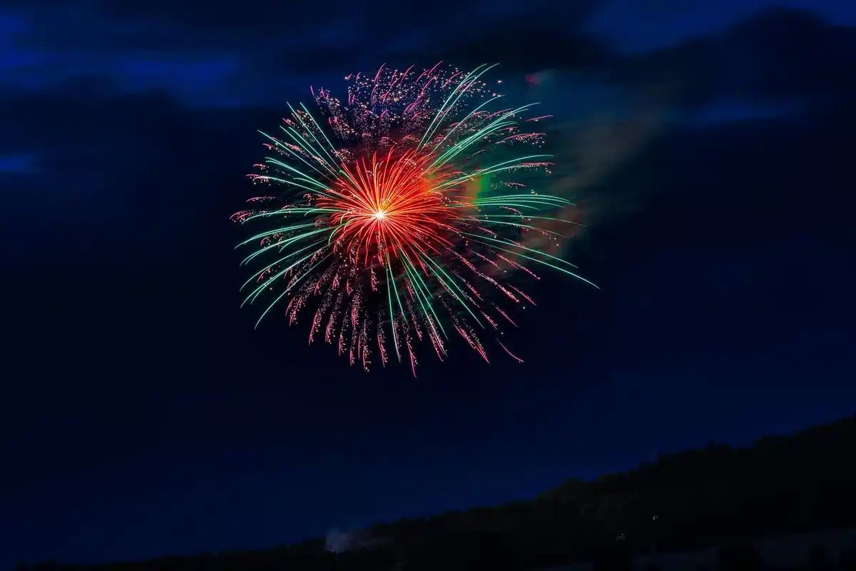 Fireworks exploding over a historic monument at night, red, white and blue colors reflected on water below, crowds silhouetted against the display, patriotic atmosphere