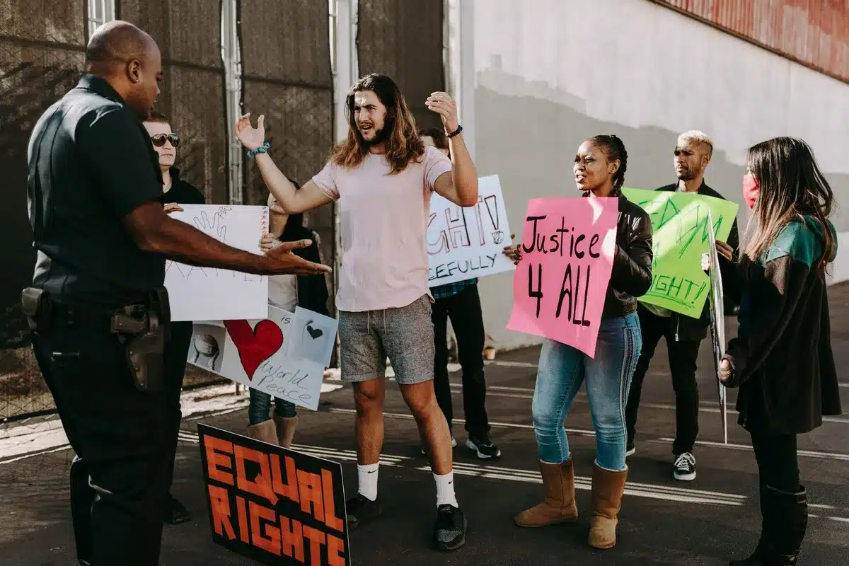 A diverse group of anonymous citizens gathered outdoors, some holding signs expressing concern about federal immigration enforcement tactics, faces distant and blurred, tension visible in body language, overcast lighting
