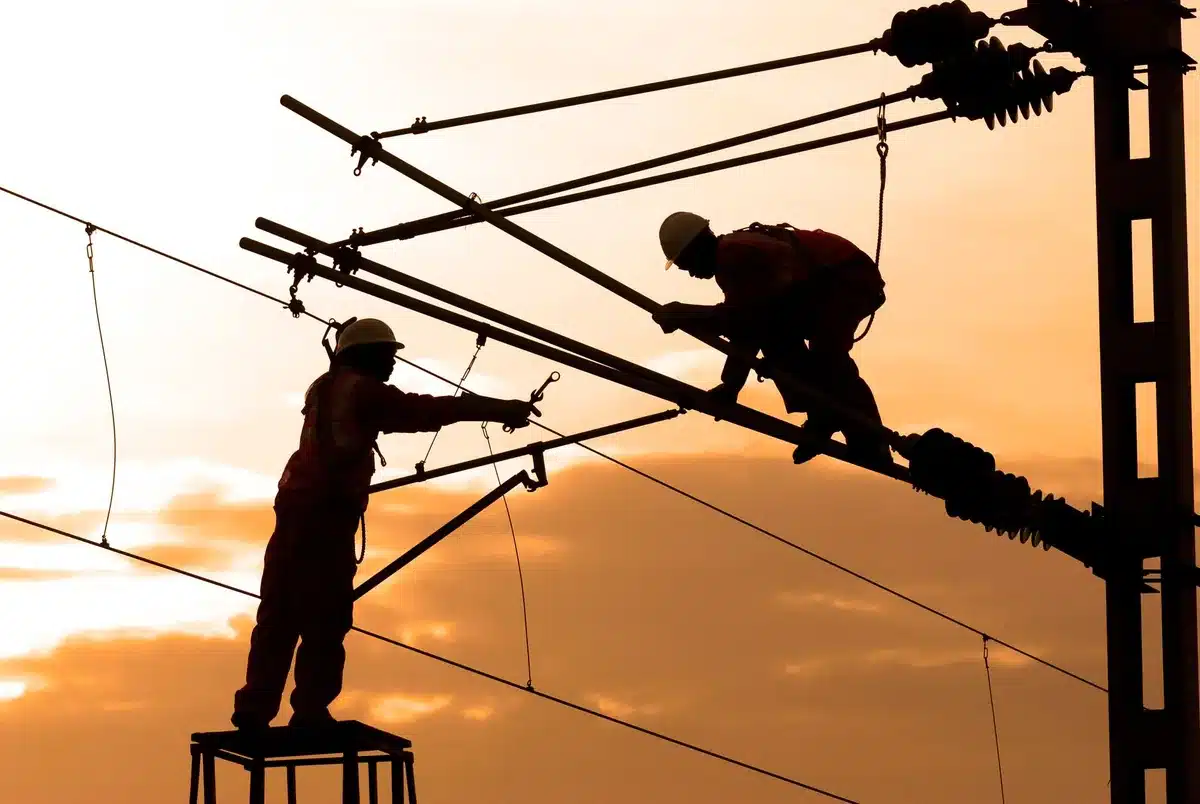 A utility truck with linemen working on damaged power poles against a hazy sky, equipment and tools visible, the scale of infrastructure damage evident in the background